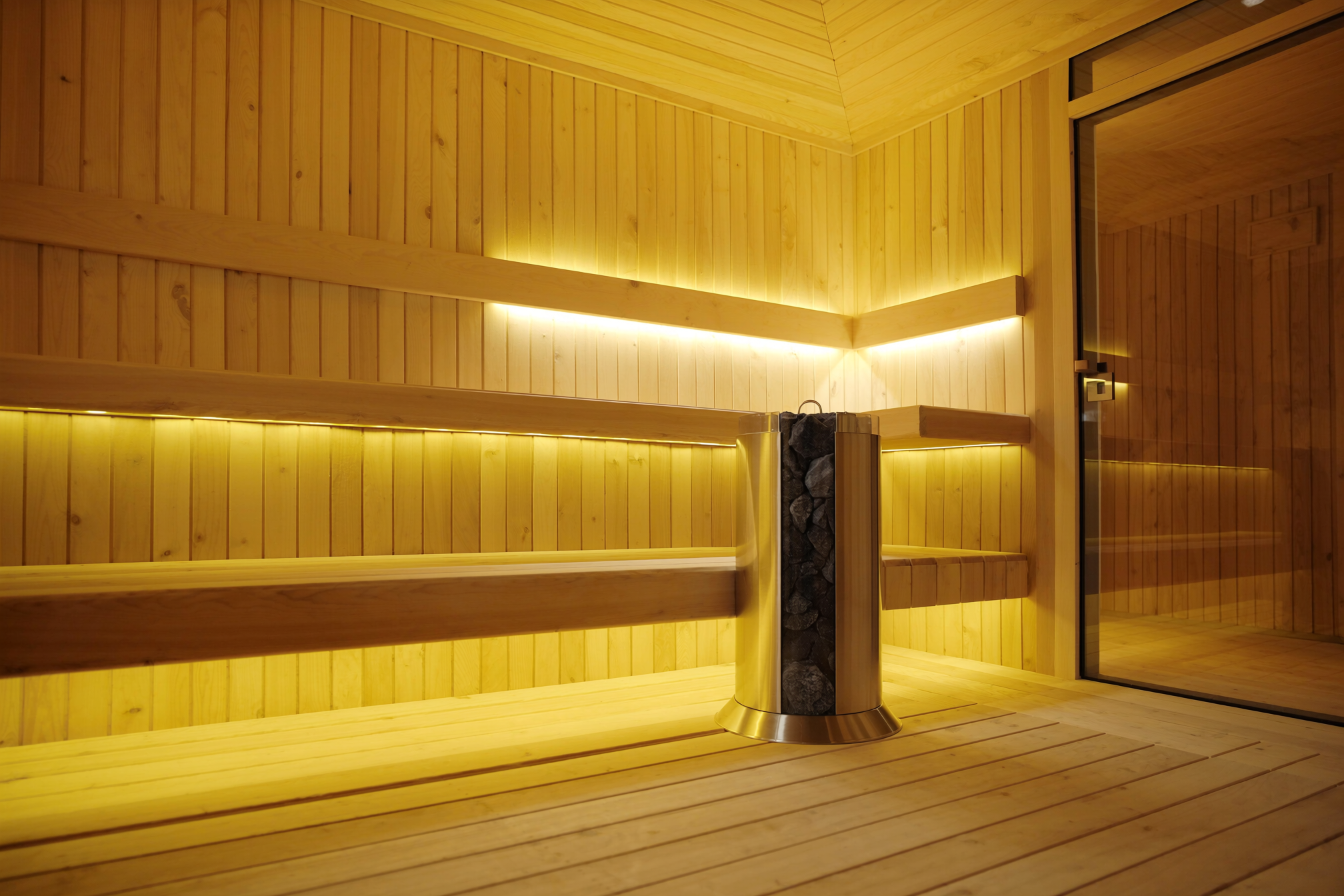 Interior of a home sauna showing light Nordic spruce cladding, two-tier bench, stainless steel electric heater with lava stones and LED strip lighting
