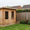 A compact timber cabin sauna on a paved base in a UK suburban back garden, near a timber boundary fence